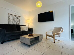 Una sala de estar moderna con un sofá oscuro, una mesa de centro de madera, un sillón de color claro, un televisor montado en la pared y una lámpara colgante.