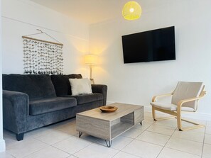 Ein modernes Wohnzimmer mit grauem Sofa, einem Holztisch, einem hellen Sessel, einem an der Wand montierten Fernseher und einem Wandbild.