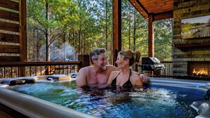 A couple in a hot tub with a wooden deck and a stone fireplace in a cabin setting. - Couples Retreat: Sauna, Massage Chairs, Hot Tub (Broken Bow)