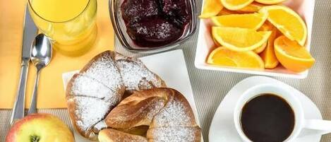 A breakfast setting with a glass of orange juice, a bowl of fruit compote, sliced oranges, a pastry, an apple, and a cup of coffee.