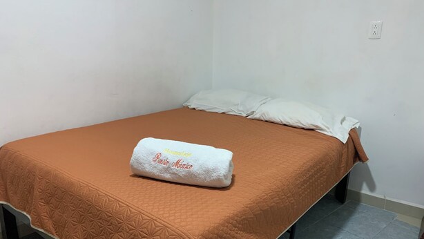 Ein Einzelbett mit einer braunen gesteppten Bettdecke und einem weißen Kopfkissen. Ein weißes Handtuch mit dem Aufdruck "Hospedaje Puero Aguas" liegt auf dem Bett.