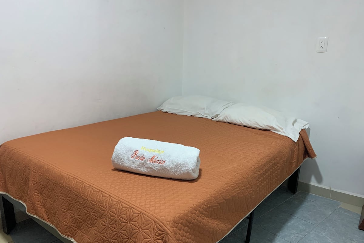Un lit simple avec une couverture matelassée marron et un oreiller blanc. Une serviette blanche avec l’inscription « Hospedaje Puero Aguas » est posée sur le lit.