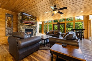 A cozy cabin interior with a stone fireplace, leather sofas, and a large window overlooking greenery.