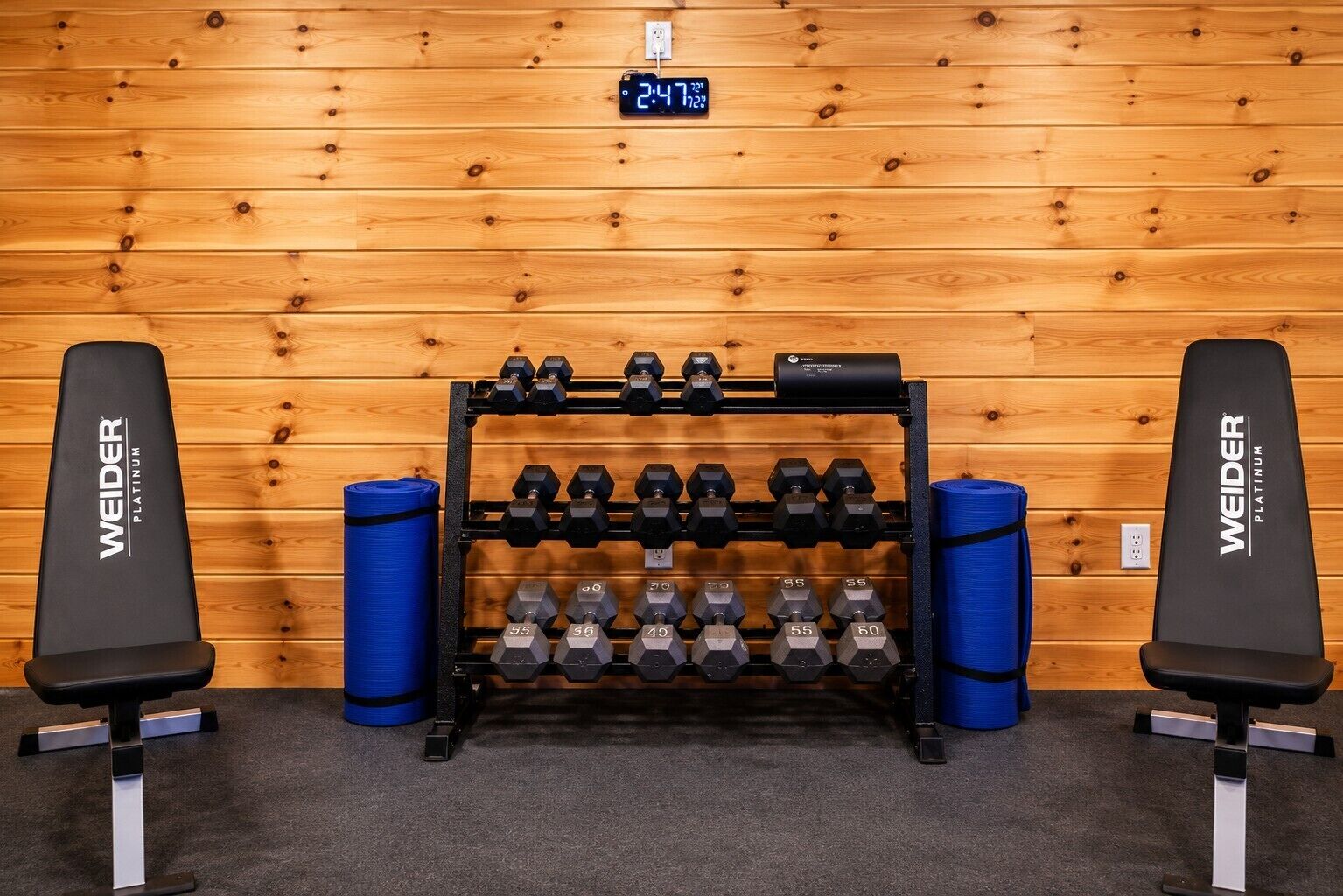 A gym with a wooden wall background, a digital clock, and dumbbells on a rack.