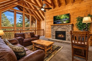 A cozy cabin interior with a stone fireplace, leather furniture, and a large window overlooking a forest. - Luxury Cabin w/ All-Inclusive Resort Amenities (Pigeon Forge)