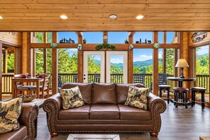 A wooden interior with a leather sofa, dining table, and chairs, large windows with a view of greenery.