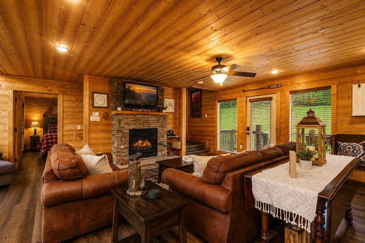 A cozy cabin interior with a stone fireplace, leather sofas, a wooden ceiling, and a television mounted above it.