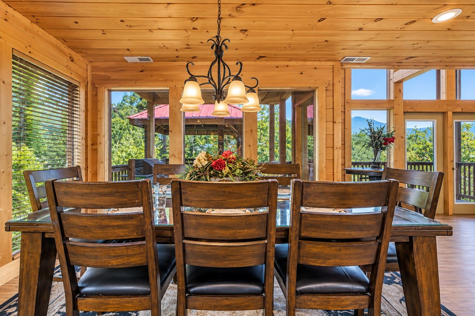 A wooden dining area with a table and chairs, a chandelier, and large windows with a view of greenery.