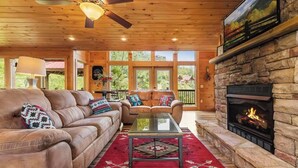 A cozy living room with a leather sofa, a glass coffee table, and a stone fireplace. - Cabin + FREE Resort Amenities | Pigeon Forge (Pigeon Forge)