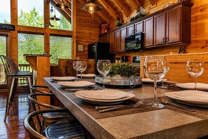 A wooden interior with a dining table set for four, kitchen cabinets, and a view of greenery outside.