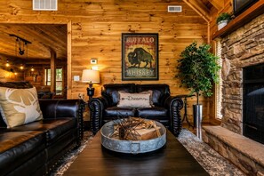 A log cabin interior with a fireplace, leather sofas, a round coffee table, and a buffalo whiskey sign.