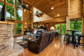 A spacious wooden cabin interior with a large living area, stone fireplace, and large windows offering views of the surrounding forest.