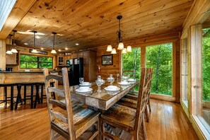 A wooden interior with a dining table set for four, a kitchen area, and large windows offering a view of greenery.