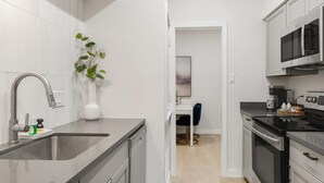 A modern kitchen with stainless steel appliances, a white countertop, and a small dining area with a blue chair. - Orchid: Studio Apt, 4 Minute Walk to A&M (College Station)