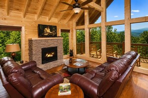 A cozy cabin interior with a fireplace, leather sofas, a television, and a balcony with a scenic view.