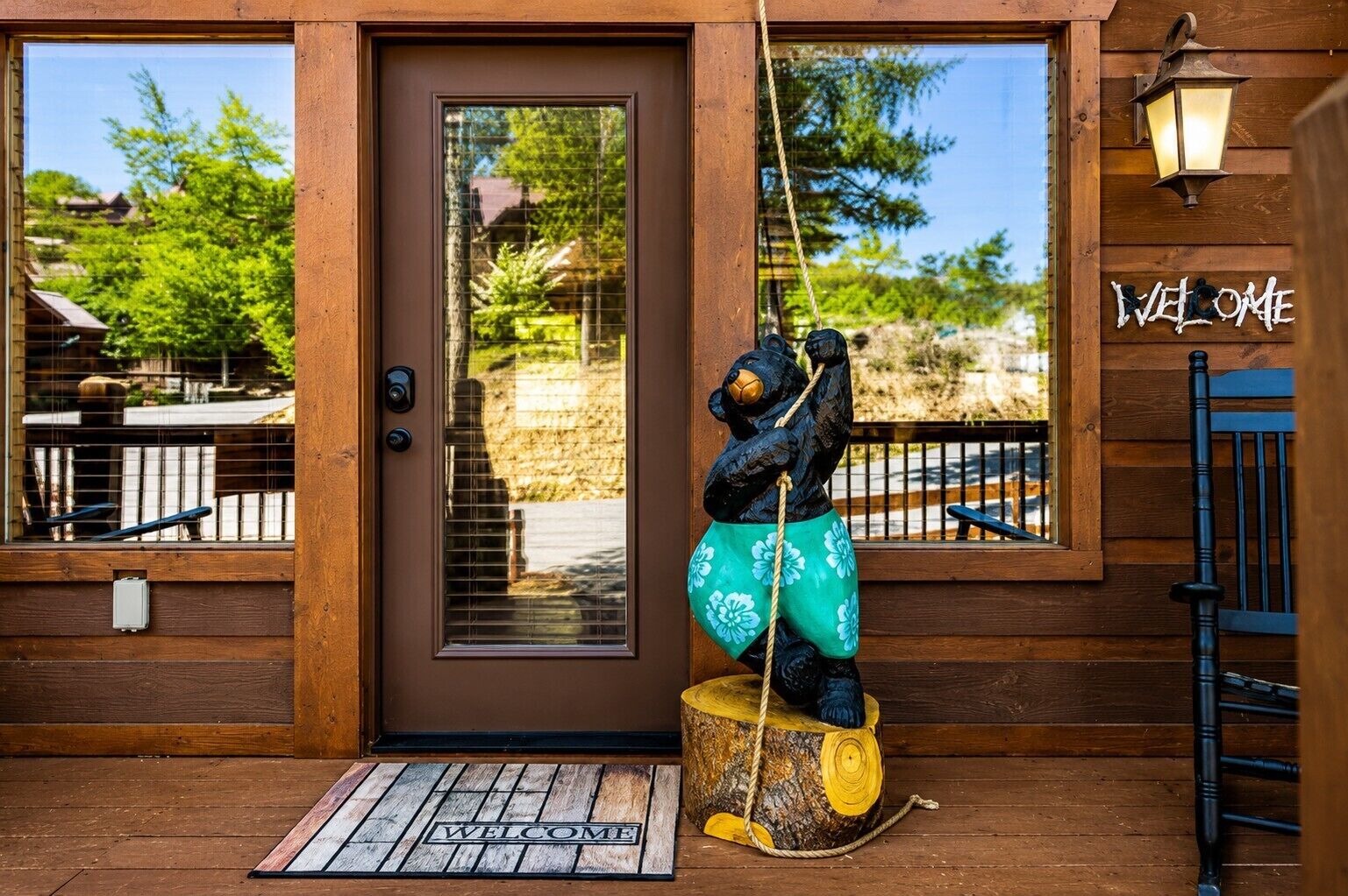 A wooden building entrance with a 'WELCOME' sign, a bear statue on a tree stump, and a doormat.