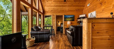 A spacious wooden cabin interior with a ceiling fan, large windows, a fireplace, and a dining area.