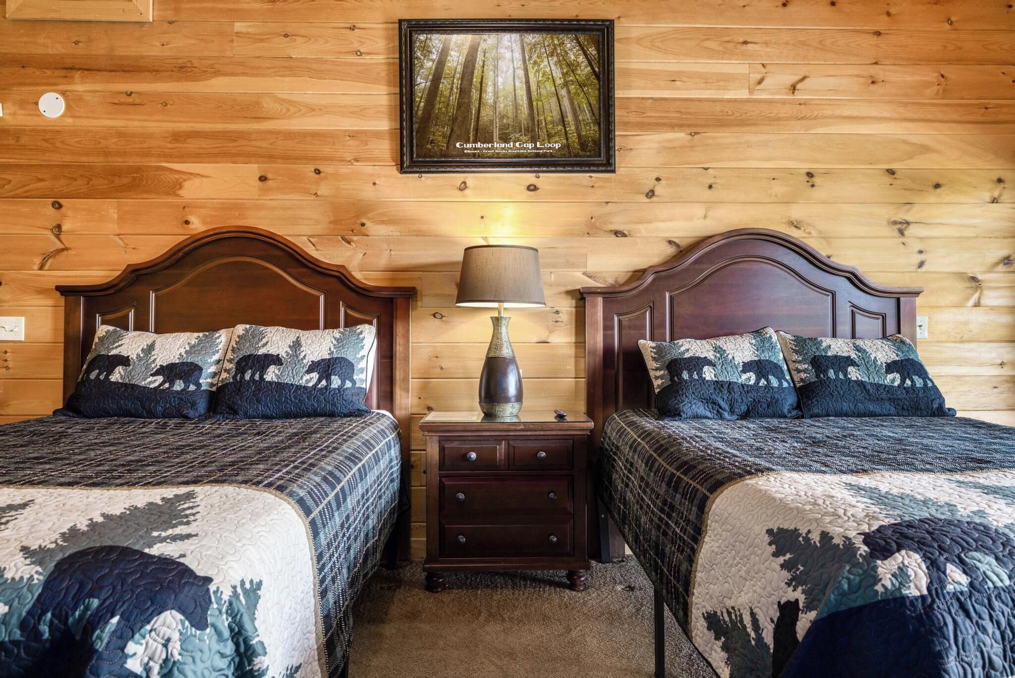 A room with two wooden beds, a nightstand with a lamp, and a framed picture on the wall.