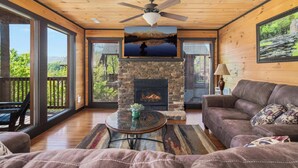 Un salon chaleureux avec une cheminée en pierre, un ventilateur de plafond et une grande fenêtre donnant sur une terrasse.