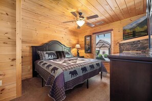 A wooden cabin bedroom with a bed, a ceiling fan, a lamp, a stone fireplace, and a window with blinds. - Cabin + FREE Resort Amenities | Pigeon Forge (Pigeon Forge)