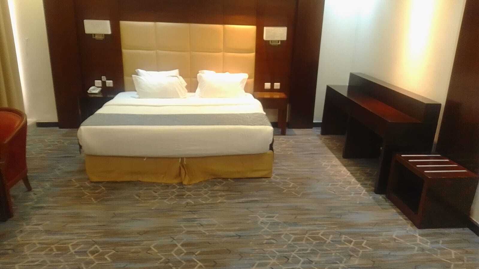 Una habitación de hotel con una cama grande, cabecera de madera, mesita de noche, un banco y suelo estampado.