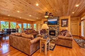 A log cabin interior with a fireplace, wooden furniture, and large windows.