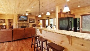 A wooden cabin interior with a kitchen area, bar stools, and a stone fireplace.