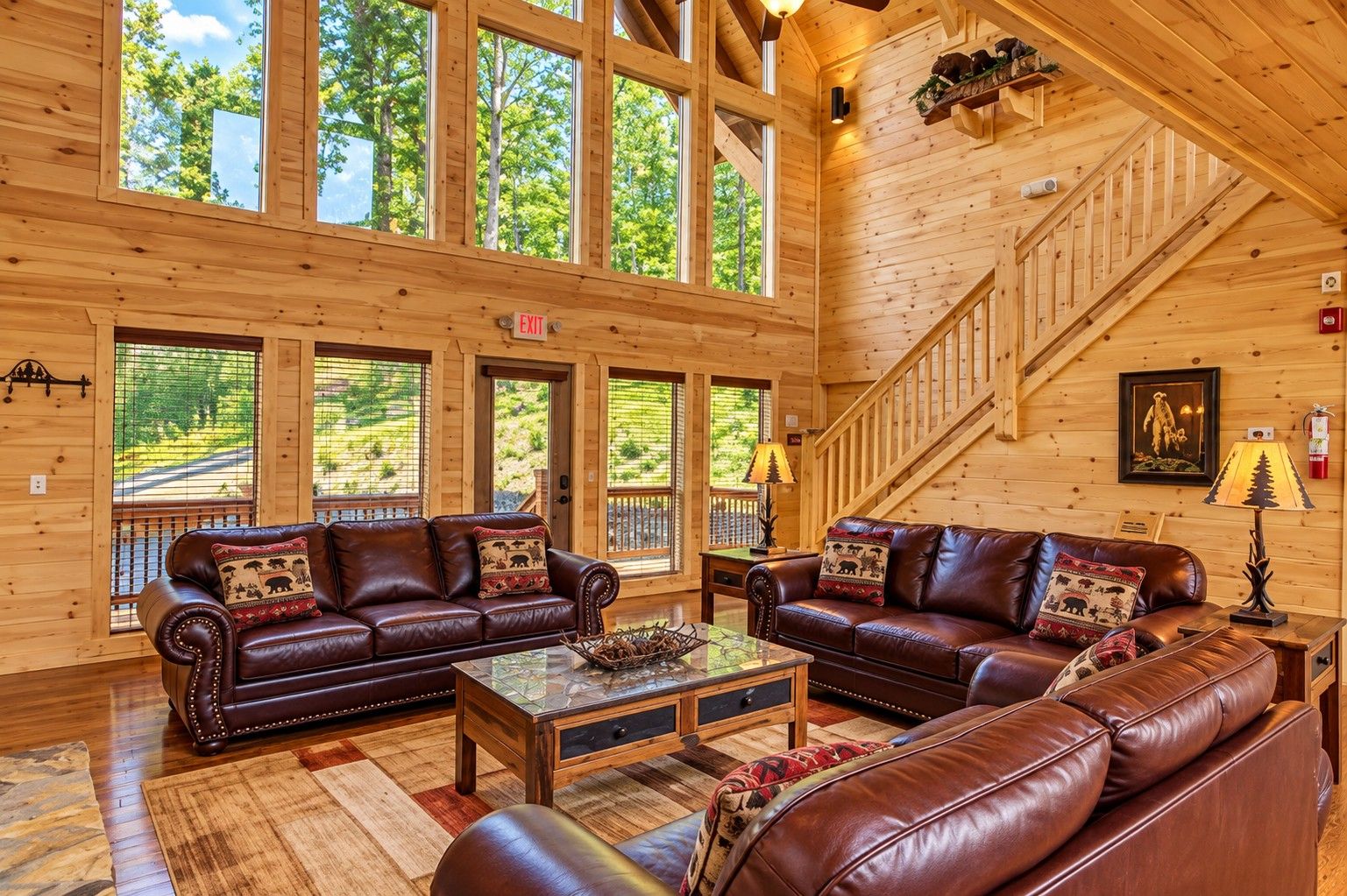 A spacious log cabin interior with leather sofas, a glass coffee table, and large windows offering a view of the outdoors.