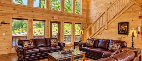A spacious log cabin interior with leather sofas, a glass coffee table, and large windows offering a view of the outdoors.