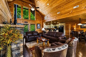 A log cabin interior with a fireplace, leather sofas, a dining area, and a kitchen.