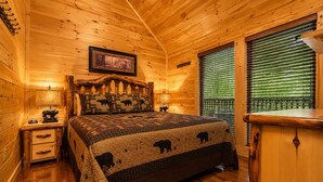 A wooden cabin bedroom with a bed, a nightstand, a dresser, a window with blinds, and a framed picture on the wall. - Cabin + FREE Resort Amenities | Pigeon Forge (Pigeon Forge)