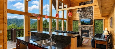 Interior de una cabaña con isla de cocina, comedor, chimenea y terraza con vistas a montañas.