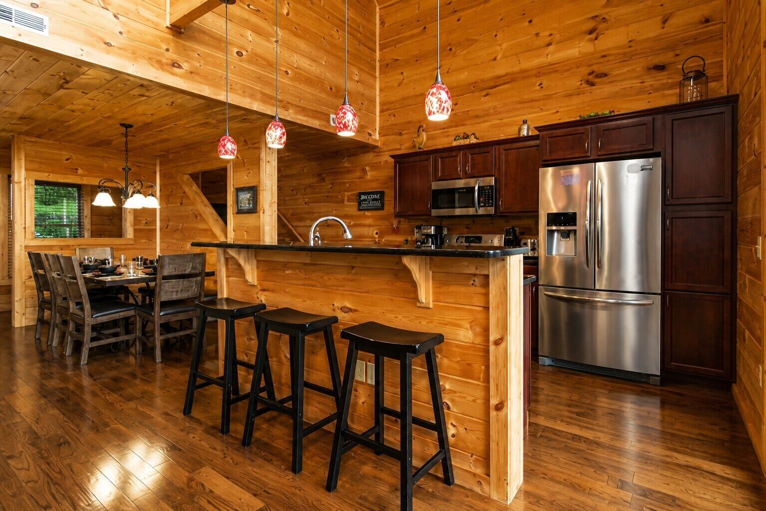 A wooden interior with a kitchen area featuring a stainless steel refrigerator, a microwave, and a sink. There are three stools at a bar counter and a dining table with chairs.
