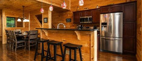 A wooden interior with a kitchen area featuring a stainless steel refrigerator, a microwave, and a sink. There are three stools at a bar counter and a dining table with chairs.