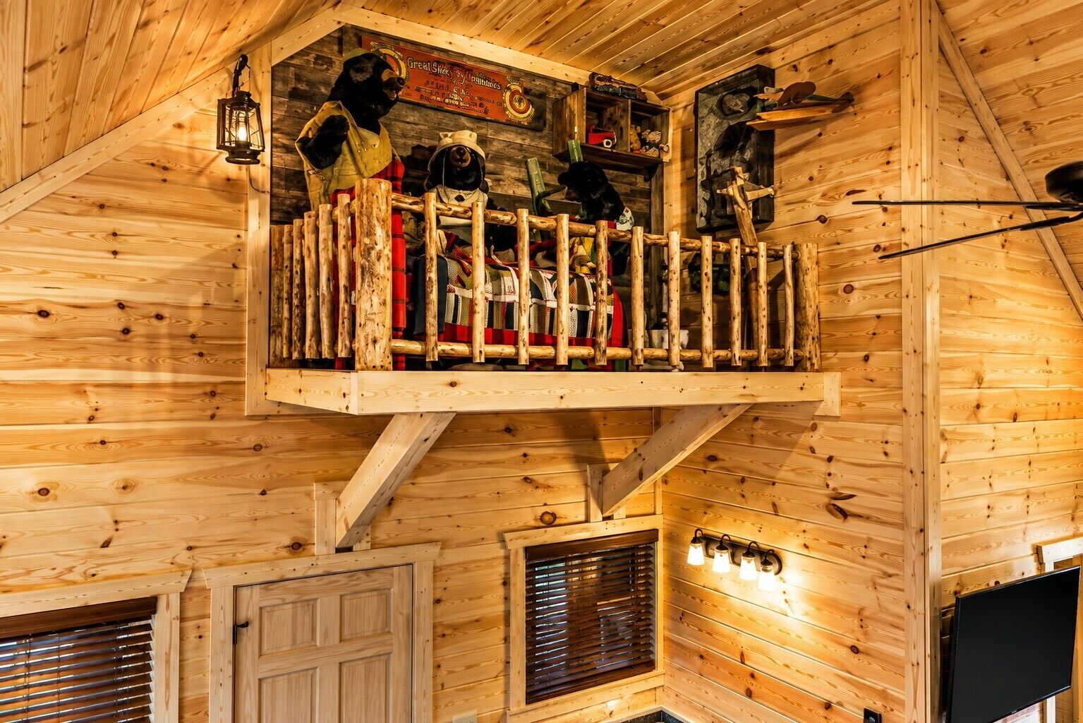 A wooden cabin interior with a lofted sleeping area, a mounted television, and a visible window with blinds.