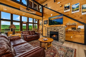 A spacious living room with a leather sofa, a fireplace, and large windows offering a view of the outdoors.