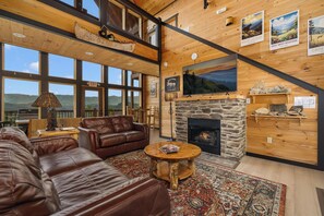 A spacious living room with a stone fireplace, leather sofas, a coffee table, and large windows offering mountain views. - Near Dollywood + Resort Amenities Included (Pigeon Forge)