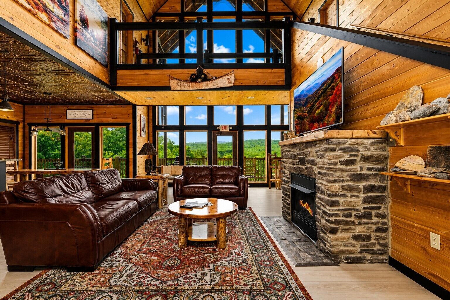 A spacious living room with a stone fireplace, leather sofas, and a large window overlooking a forested landscape.
