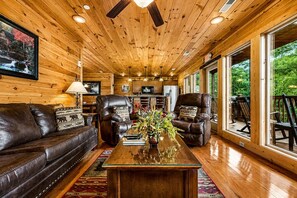 A wooden interior with a leather sofa, two recliners, a coffee table, and a kitchen area visible through glass doors.