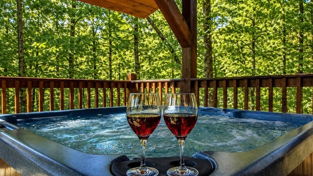 A hot tub with two glasses of red wine on a wooden deck surrounded by trees. - Cabin + FREE Resort Amenities | Pigeon Forge (Pigeon Forge)