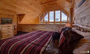 Quarto de cabana de tronco com teto inclinado, uma janela grande, uma cama com manta listrada e um aparador de madeira.