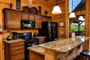 A kitchen with wooden cabinets, granite countertops, a black refrigerator, a microwave, a stove, and a coffee maker. - Cabin + FREE Resort Amenities | Pigeon Forge (Pigeon Forge)