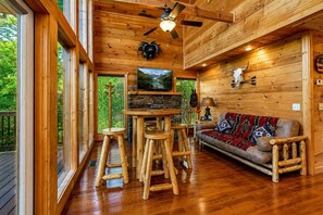 A log cabin interior with a wooden ceiling, a stone fireplace, a television, a sofa, and a wooden bar stool.