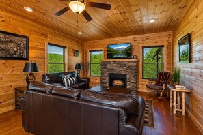 A log cabin interior with a fireplace, leather sofa, television, and a ceiling fan. - Cabin + FREE Resort Amenities | Pigeon Forge (Pigeon Forge)