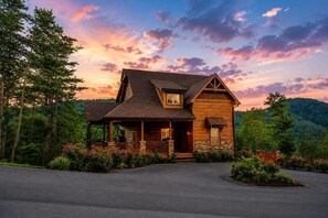 A log cabin with a porch, surrounded by lush greenery and a mountain backdrop. - Luxury Cabin w/ All-Inclusive Resort Amenities (Pigeon Forge)