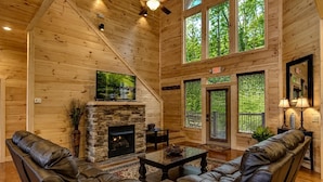 A log cabin interior with a stone fireplace, leather sofas, a television, and large windows.