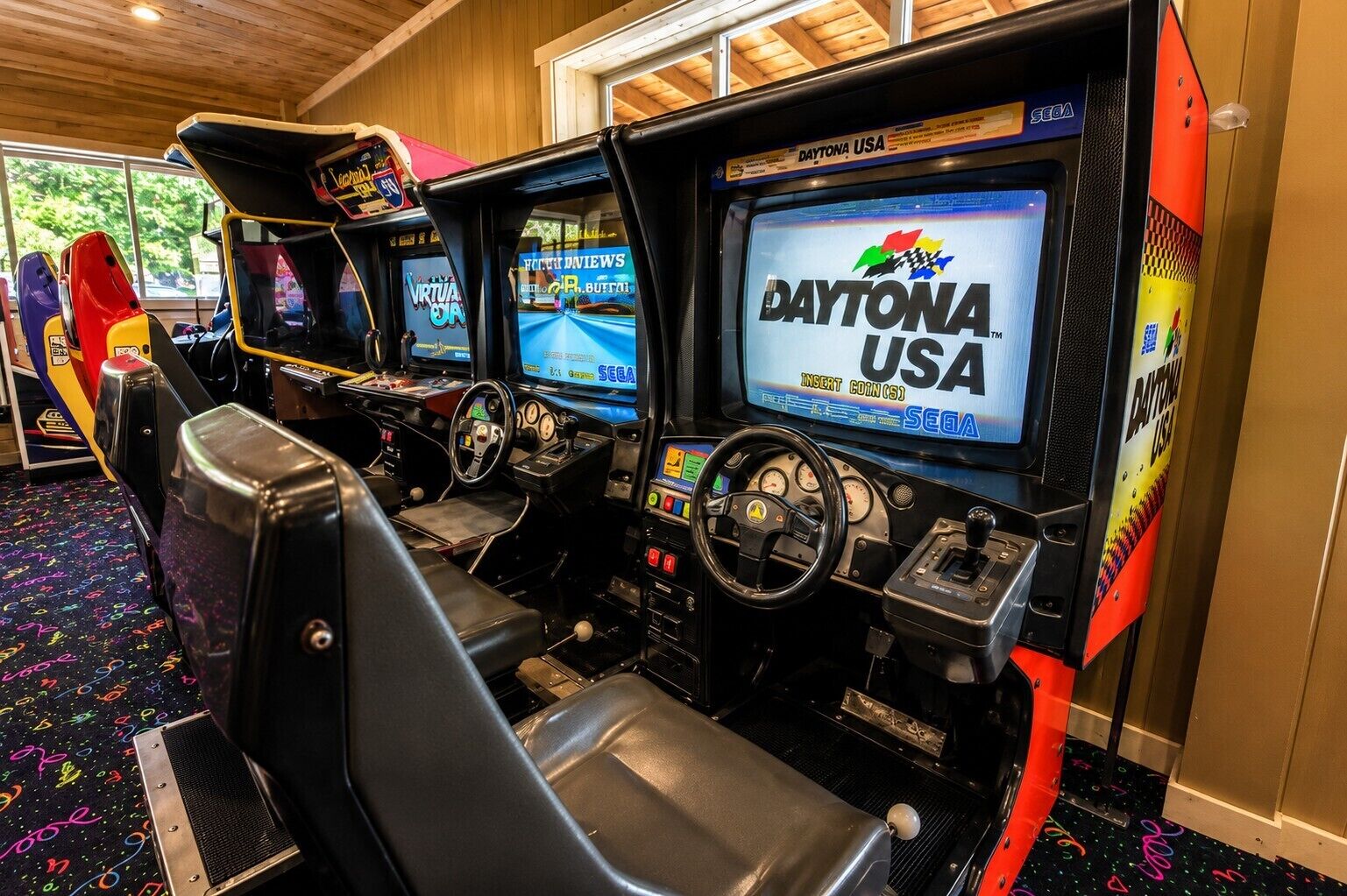 A virtual racing game arcade with multiple racing simulators, each featuring a steering wheel and a monitor displaying the Daytona USA logo.