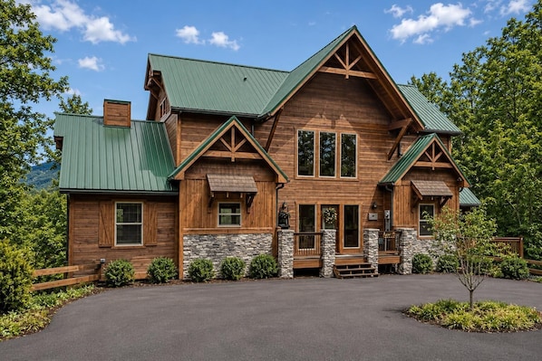 A large log cabin with a stone foundation, multiple windows, and a green metal roof. - Cabin + FREE Resort Amenities | Pigeon Forge (Pigeon Forge)