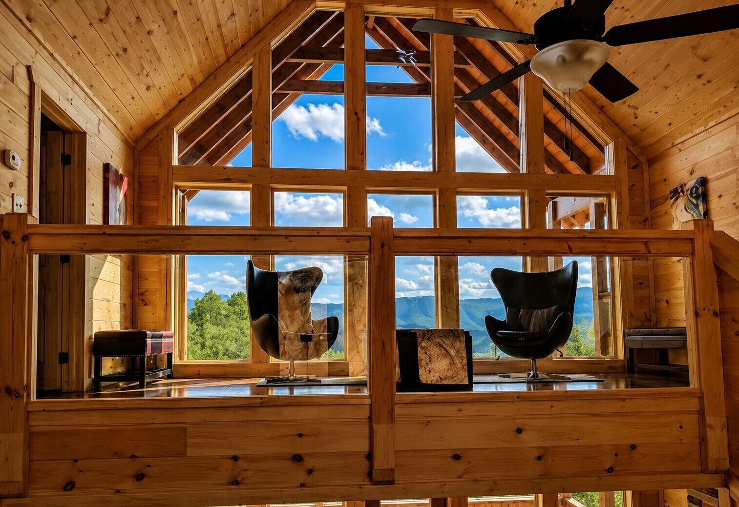 A wooden cabin interior with large windows, a ceiling fan, and two black chairs.