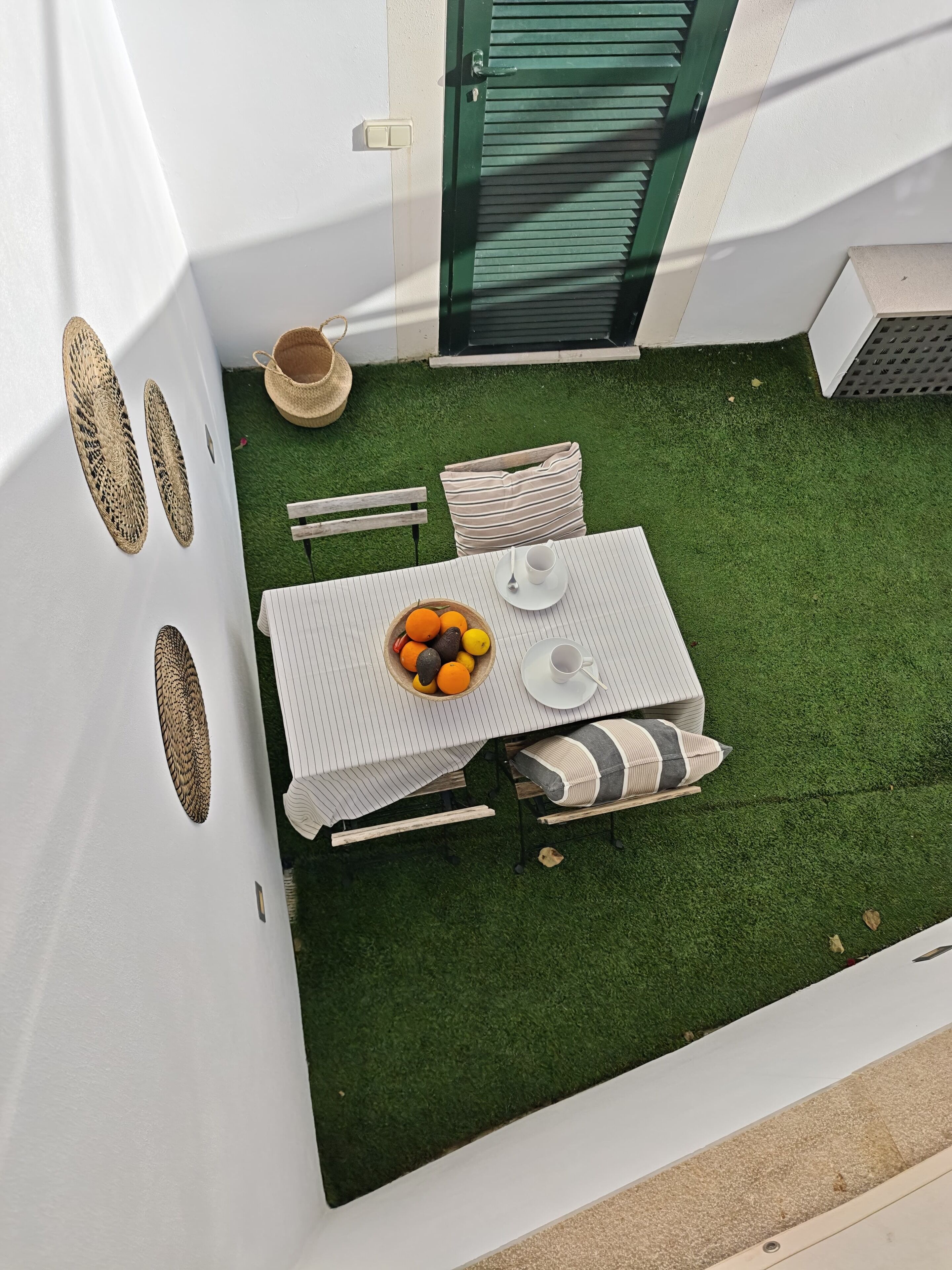 Un pequeño espacio exterior para comer con una mesa preparada para dos personas, rodeado de césped artificial, una cesta de mimbre y un banco de madera.